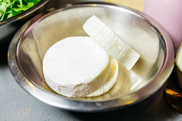Whole and Sliced Wheel of Brie Cheese. A whole wheel of Brie cheese alongside a sliced piece on a metal bowl, ready for a gourmet meal or snack preparation.