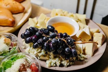 Cheese Platter with Grapes and Honey Dip. A variety of gourmet cheeses accompanied by fresh black grapes and a bowl of honey, perfect for a cheese board setup or a wine pairing event.