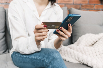 Unrecognizable woman sitting on couch, holding credit card and using mobile phone, smartphone for online shopping or internet payment