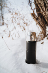 Black thermos bottle with a silver lid sits in the snow near a tree trunk on a sunny winter day. - obrazy, fototapety, plakaty