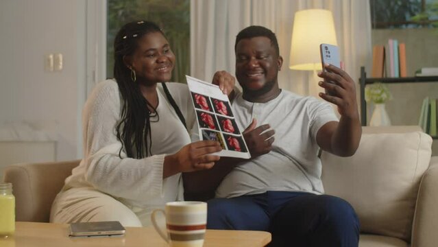 Medium long shot of African American young couple showing ultrasound image of their unborn baby via video call on smartphone
