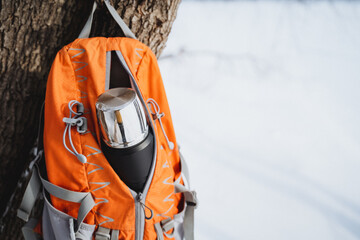 Orange backpack with silver thermos bottle hanging on tree in winter forest