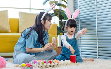 Asian cute little children girl wearing funny bunny ears headbands and young pretty mother smile decorating painting eggs while sitting on floor in living room, Family preparing for Easter holiday.