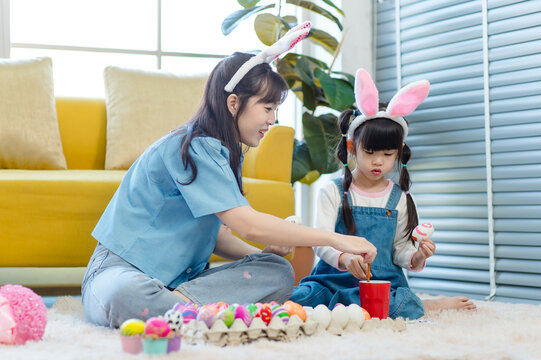 Asian cute little children girl wearing funny bunny ears headbands and young pretty mother smile decorating painting eggs while sitting on floor in living room, Family preparing for Easter holiday.
