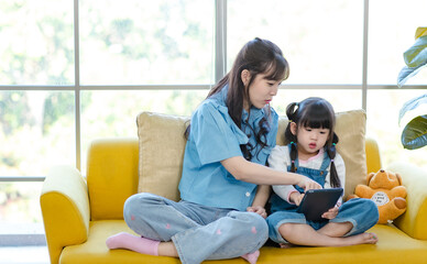 Asian young mother and her little cute daughter girl learning study from tablet computer, sitting on yellow sofa in living room at home, Happy family, Education and early development concept.