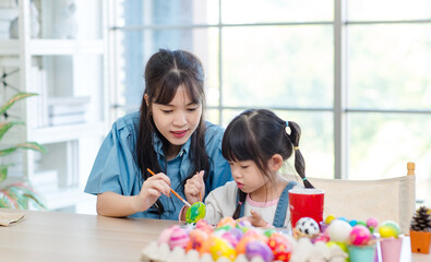 Obraz na płótnie Canvas Asian cute little children girl wearing funny bunny ears headbands and young happy mother smile decorating painting eggs while sitting together in living room table family preparing for Easter holiday