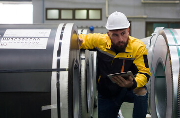A man wearing a safety helmet holding tablet computer inspects large metal coils at an industrial...