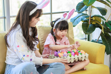 Obraz na płótnie Canvas Happy asian cute little children girl wearing bunny ears headbands and young happy mother smile, play with eggs while sitting together on sofa in living room during preparation for Easter holiday