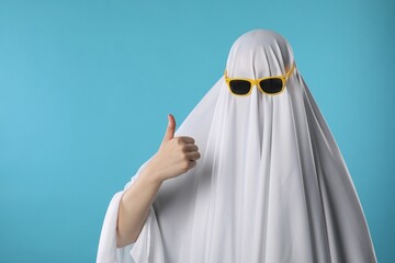 Stylish ghost. Woman covered with white sheet in sunglasses showing thumbs up on light blue...
