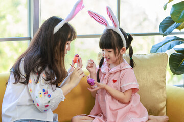 Obraz premium Asian cute little children girl wearing funny bunny ears headbands and young happy mother smile decorating painting eggs while sitting together on sofa in living room, Family preparing Easter holiday.