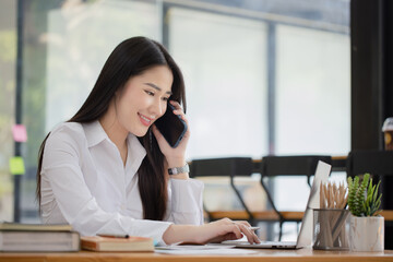 Happy asian young businesswoman using digital smartphone sitting in office working space, Asian female employee using laptop talking on the phone at workplace.