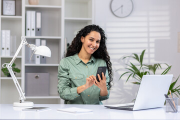 Professional hispanic woman working in a bright office space with a laptop and mobile phone,...