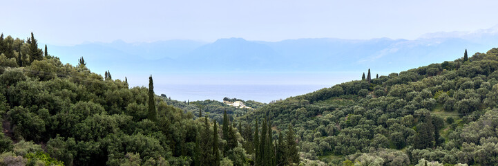 Fototapeta na wymiar Green hills on the seashore and the silhouettes of the mountains on the horizon. View from the Greek island of Corfu to the mountains on the coast of Albania