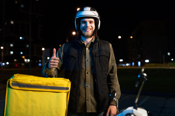 A man in a helmet near a scooter in the evening. The deliveryman during the night shift