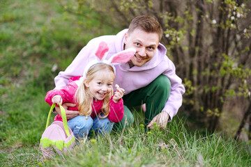 Caucasian father and daughter with bunny ears collecting Easter eggs and putting Easter eggs in baskets in the park. Easter egg hunt concept