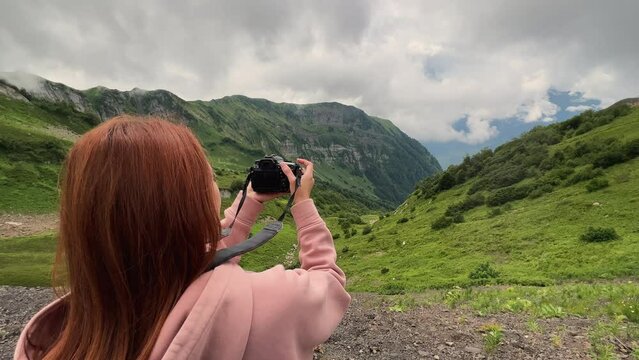 Beautiful red-haired girl in pink hoodie photographing mountain landscape on a summer warm day in slowmotion