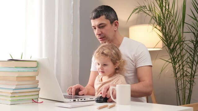 Positive family spending free time with computer entertainment man dressed casual T-shirt using laptop with his toddler baby sitting at table at home playing video games