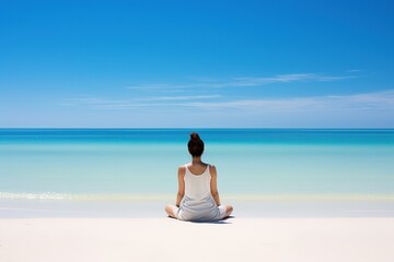 Fototapeta na wymiar A woman in white summer clothes sits, viewed from the back, in the lotus position on the ocean shore and meditates while looking into the distance. White sandy beach, blue sky with light clouds.