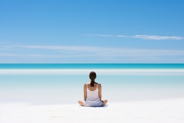 Fototapeta na wymiar A woman in white summer clothes sits, viewed from the back, in the lotus position on the ocean shore and meditates while looking into the distance. White sandy beach, blue sky with light clouds.