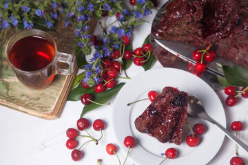 Chocolate cake with sweet cherry, cup of tea on vintage book and branch of blue flowers on white background..