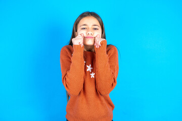 Pleased Young beautiful teen girl wearing orange knitted sweater with closed eyes keeps hands near...