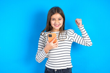 Young beautiful teen girl wearing striped T-shirt  holding in hands cell and rising his fist up being excited after reading good news.