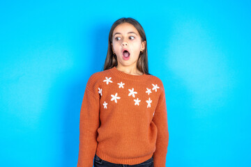 Shocked Young beautiful teen girl wearing orange knitted sweater stares bugged eyes keeps mouth...
