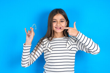 Young beautiful teen girl wearing striped T-shirt holding an invisible aligner and pointing perfect straight teeth. Dental healthcare and confidence concept.