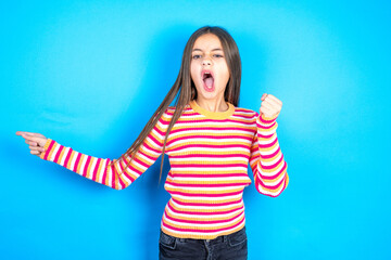 Young kid girl wearing striped t-shirt points at empty space holding fist up, winner gesture.