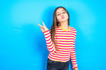 Young kid girl wearing striped t-shirt makes peace gesture keeps lips folded shows v sign. Body...
