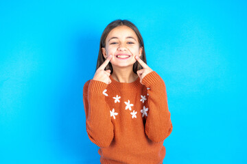 Happy Young kid girl wearing orange sweater with toothy smile, keeps index fingers near mouth, fingers pointing and forcing cheerful smile