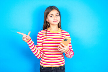 Astonished Young kid girl wearing striped t-shirt holding her telephone and pointing with finger...