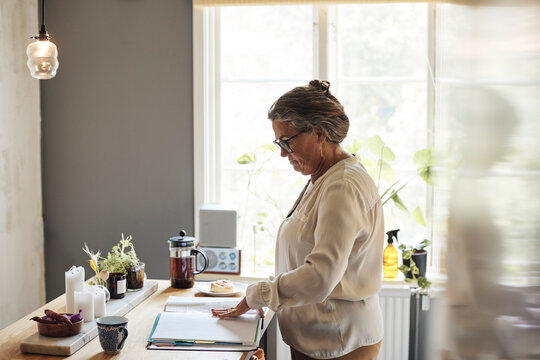 Side view of woman examining financial bills while standing in kitchen at home