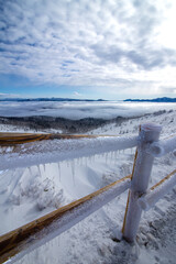 雪に覆われた峠の頂上の朝。木の柵は凍り付いている。青空には幅広い雲が広がって朝陽を遮り、眼下には雲海が見える。日本の北海道の美幌峠。寒さの厳しい冬の穏やかな晴れの日。