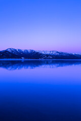 淡い紫色のグラデーションに染まる夜明けの空と雪山を水面に反射する湖。日本の北海道の旅行地である屈斜路湖。雄大で美しい自然。