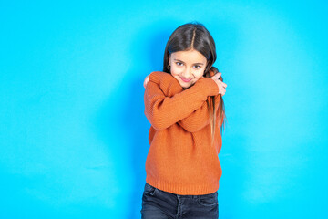 Charming pleased beautiful kid girl wearing orange sweater embraces own body, pleasantly feels...