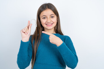 beautiful kid girl wearing blue sweater holding an invisible aligner and pointing at it. Dental healthcare and confidence concept.