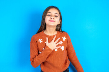 Joyful beautiful kid girl wearing orange sweater expresses positive emotions recalls something funny keeps hand on chest and giggles happily. - obrazy, fototapety, plakaty