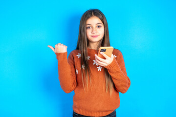 Young kid girl wearing orange knitted sweater points thumb away and shows blank space aside, holds...