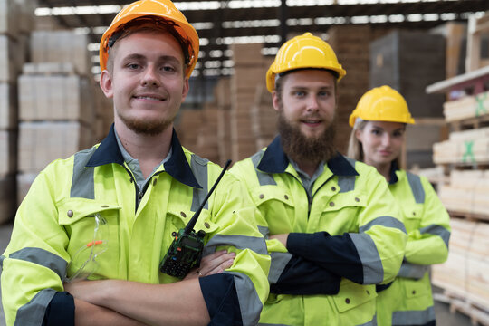 Group of male and female warehouse workers standing together with crossed arms and smiling in industry factory, wearing safety uniform and helmet. Warehouse workers working in warehouse storage
