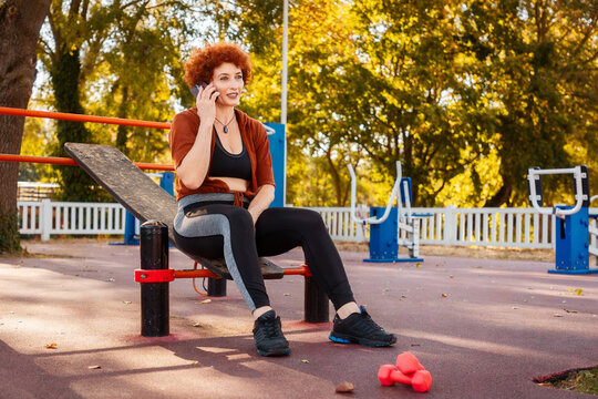 Mid adult happy Caucasian curly-haired woman sitting at sports ground and call with cell phone. Concept of motivation and activity