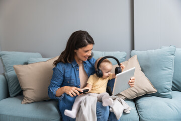 Young mother trying to entertain her newborn baby by playing cartoons and funny videos on digital tablet while sitting on sofa at home. Woman using internet for fun with her little child.