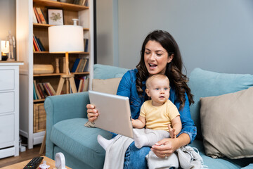 Young mother trying to entertain her newborn baby by playing cartoons and funny videos on digital tablet while sitting on sofa at home. Woman using internet for fun with her little child.