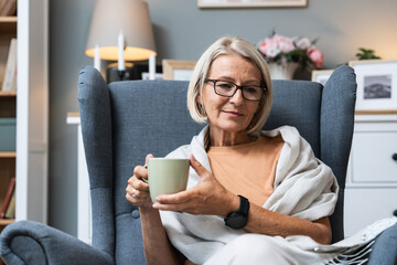 Simple living. Senior woman sitting alone on chair at home drinking tea or coffee enjoying her time...