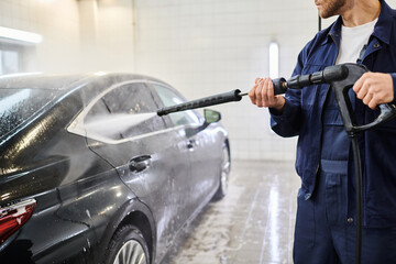 cropped view of devoted professional worker in blue uniform using hose to wash black modern car