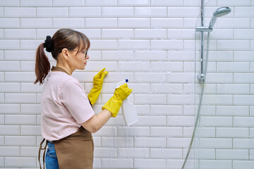 Middle-aged woman in an apron cleaning tiles in shower in bathroom