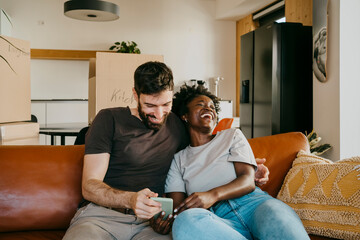Happy multiracial couple enjoying sharing smart phone while sitting on sofa in living room at home