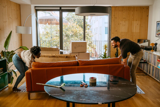 Multiracial couple helping each other while keeping sofa in living room at new home