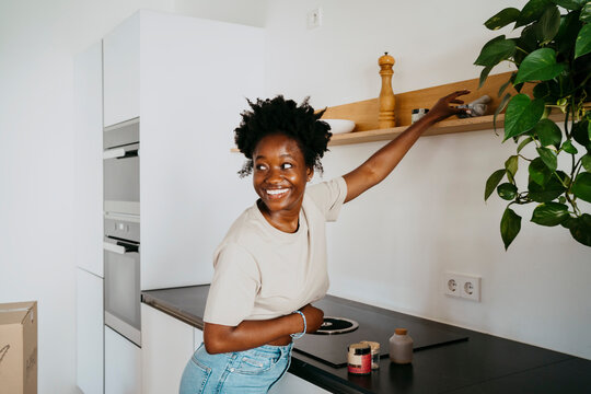 Smiling woman looking away while reaching at shelf in kitchen of new home