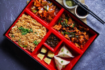 Bento box on a grunge background with wasabi, soy sauce, and chopsticks. Containing fried rice, chicken, beef, sushi, dumplings, Chili sauce, mayonnaise, food photography, social media post